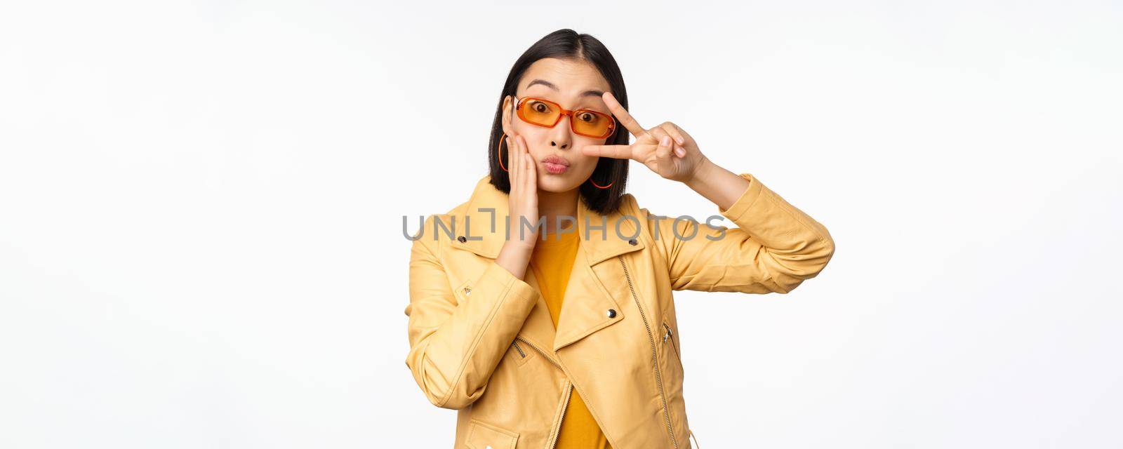 Portrait of stylish asian modern girl, wearing sunglasses and yellow jacket, showing peace, v-sign gesture, standing over white background, happy smiling face.