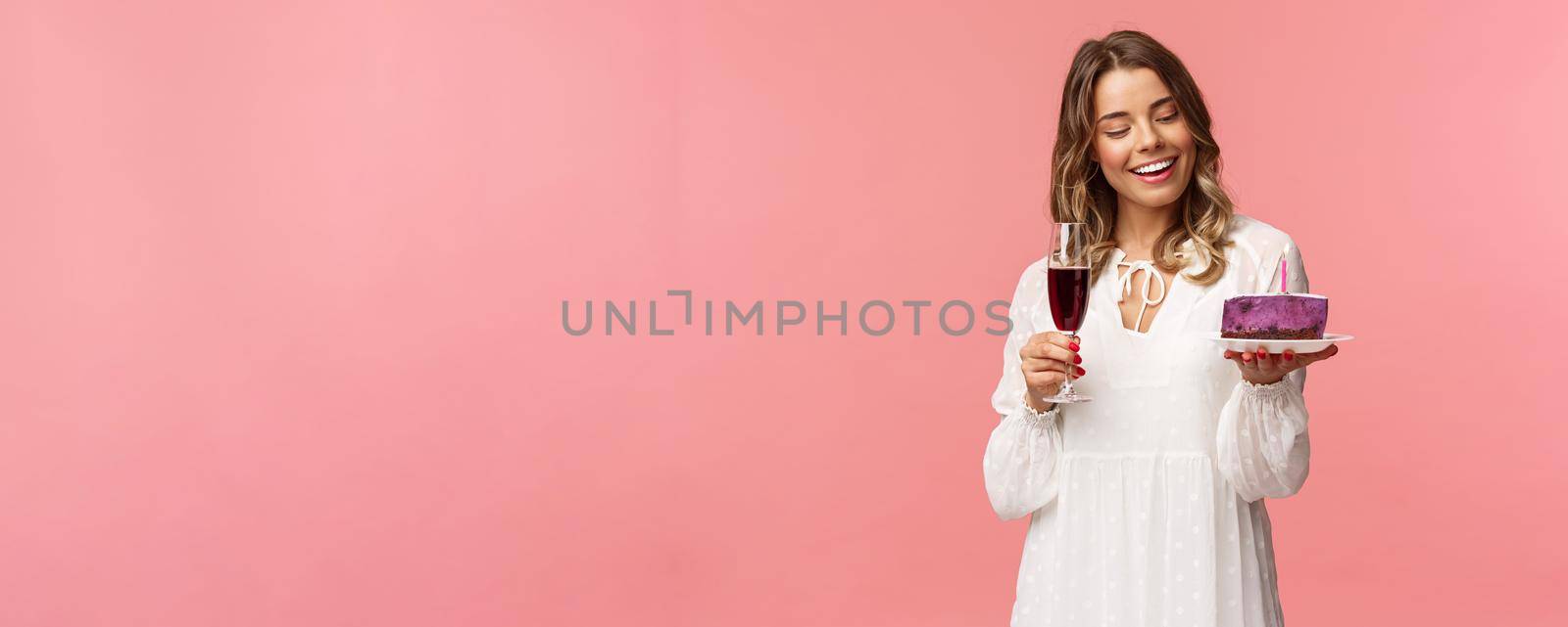 Holidays, spring and party concept. Portrait of tender, elegant young blond woman holding glass of wine and cake with lit candle, smiling pleased, celebrating birthday, pink background.
