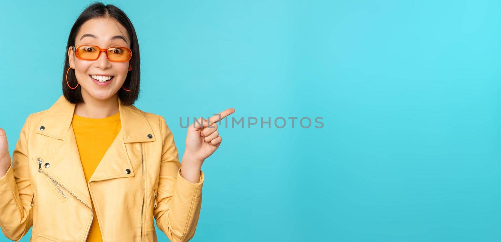 Happy stylish chinese girl in sunglasses, points fingers sideways, invites to check out, see store discount, shows left and right, stands over blue background.