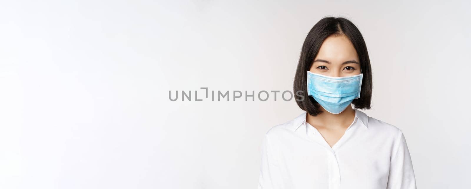 Covid and healthcare concept. Close up portrait of asian woman, office lady in face mask, smiling, using protection from coronavirus during pandemic, white background.