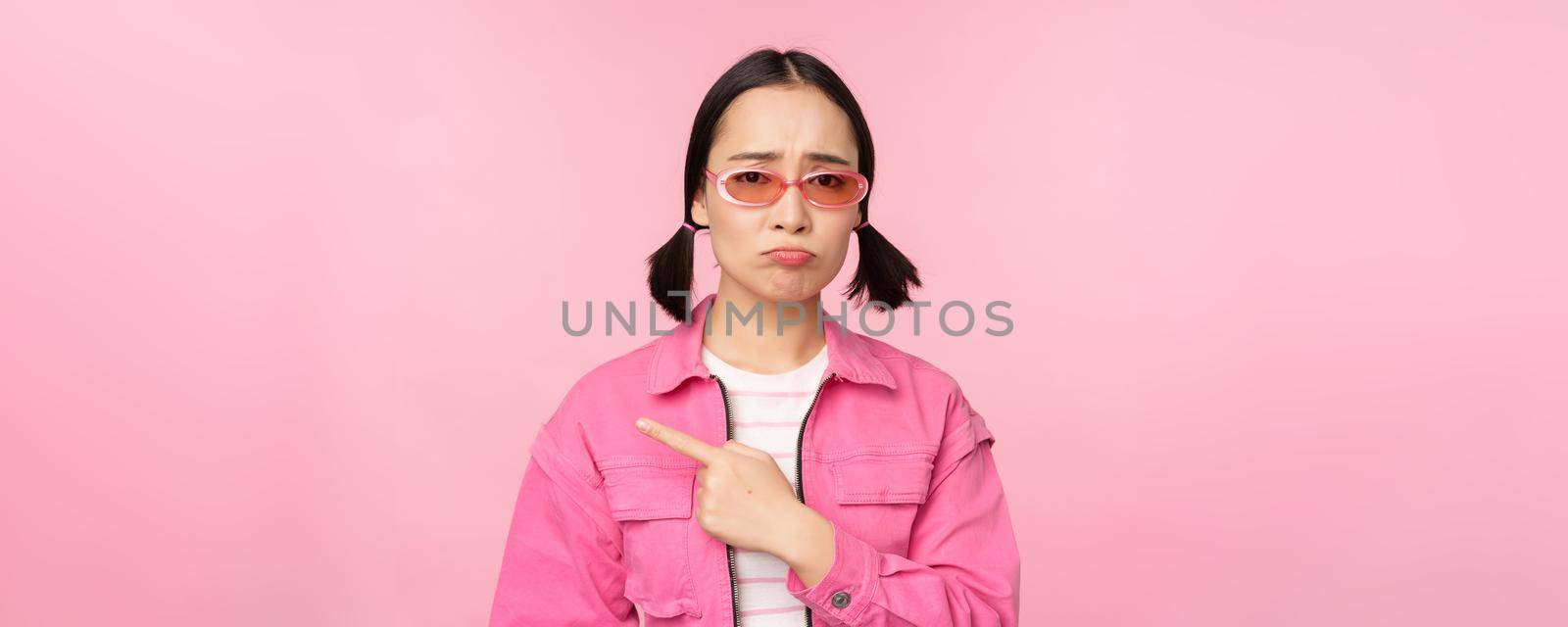 Potrait of cute korean girl in sunglasses, pointing left and looking disappointed, sulking upset, standing over pink background.