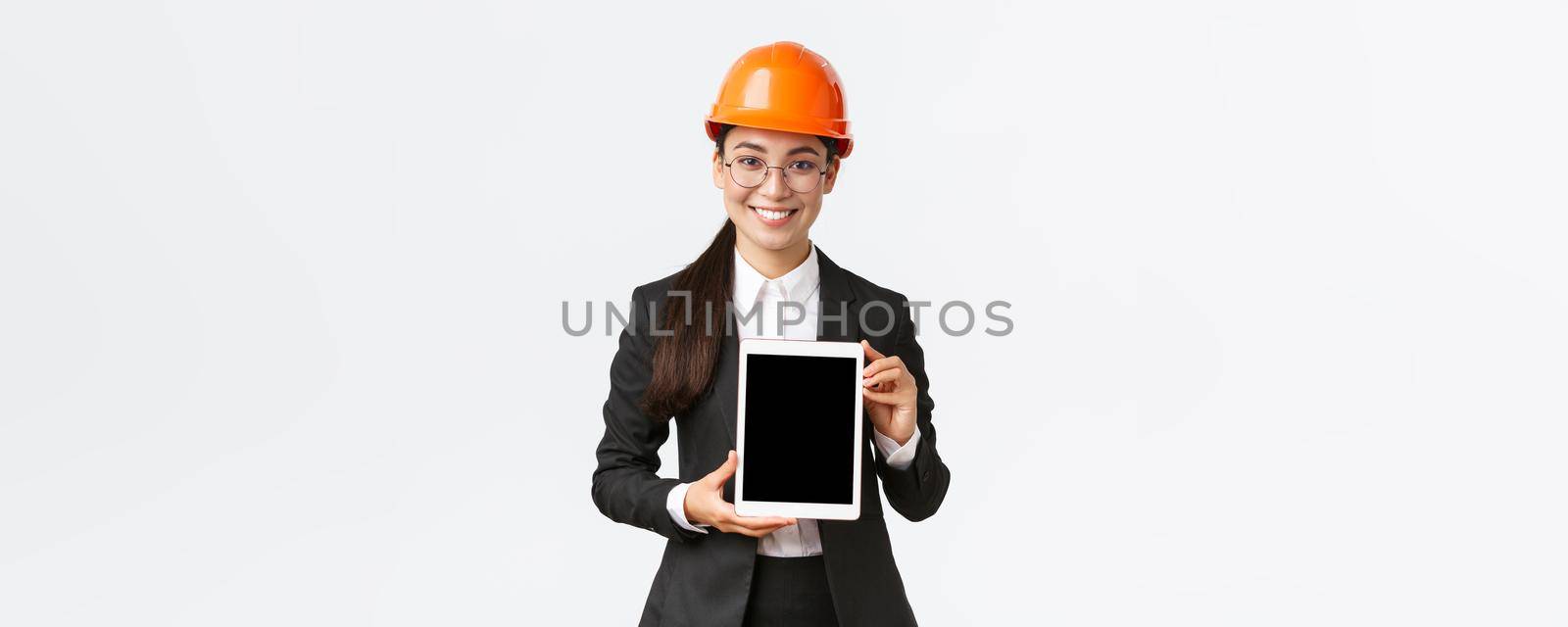 Smiling professional female engineer, saleswoman or estate agent showing location to client, standing in safety helmet and business suit, introduce construction project at tablet screen.
