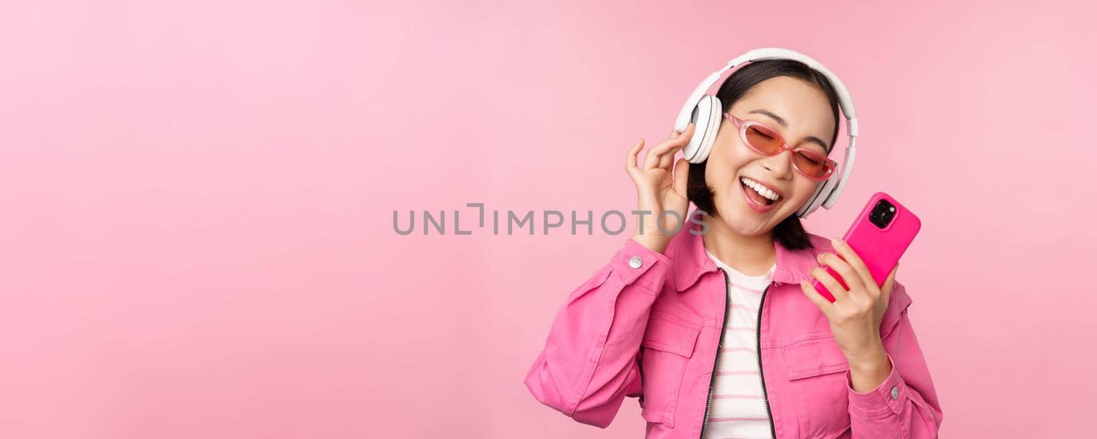 Stylish asian girl dancing with smartphone, listening music in headphones on mobile phone app, smiling and laughing, posing against pink background.