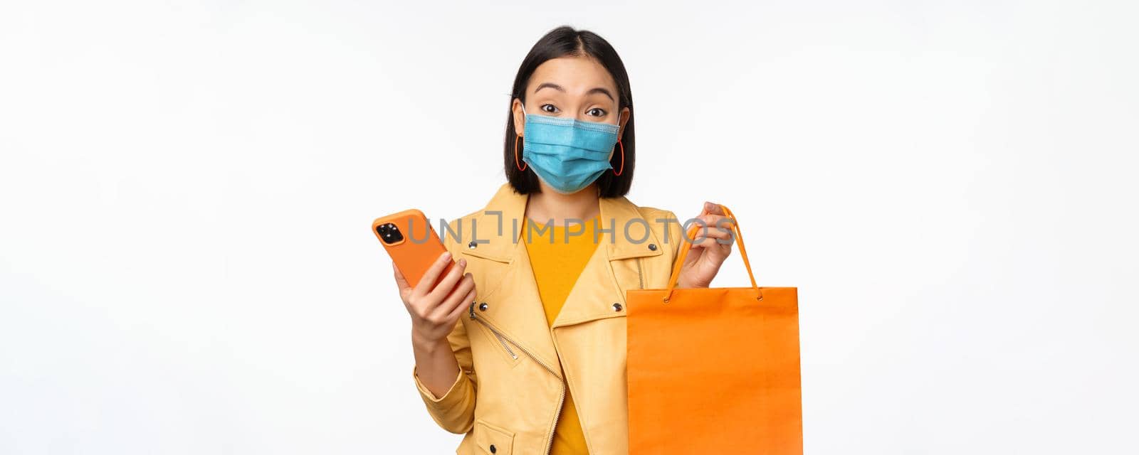 Image of stylish asian girl shopper, holding smartphone and shopping bag without store logo, wearing medical face mask from covid-19, white background.