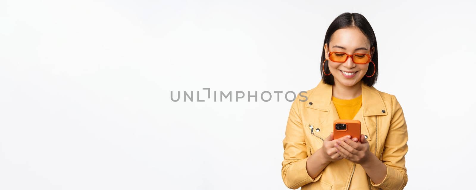 Modern asian girl in sunglasses using her mobile phone, smiling and looking happy, posing against white background. Copy space