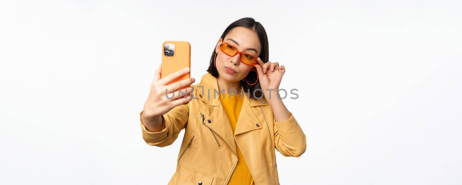 Stylish, beautiful asian girl in sunglasses, taking selfie on smartphone, posing for photo, holding mobile phone, white studio background.