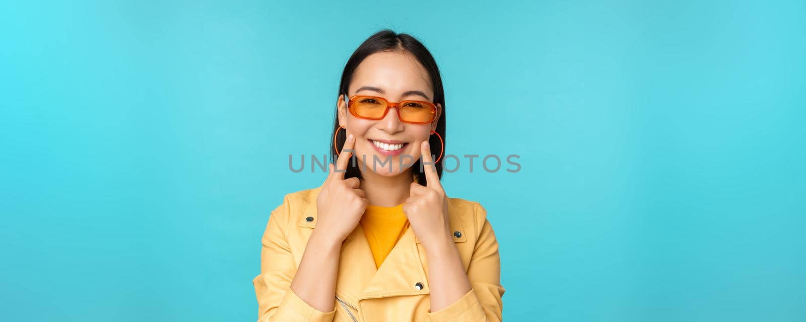 Close up portrait of asian young woman in sunglasses, smiling and looking romantic, standing happy over blue background. Copy space