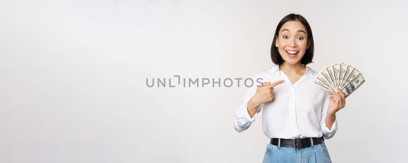 Portrait of young asian woman pointing at her money dollars, showing cash, standing over white background.
