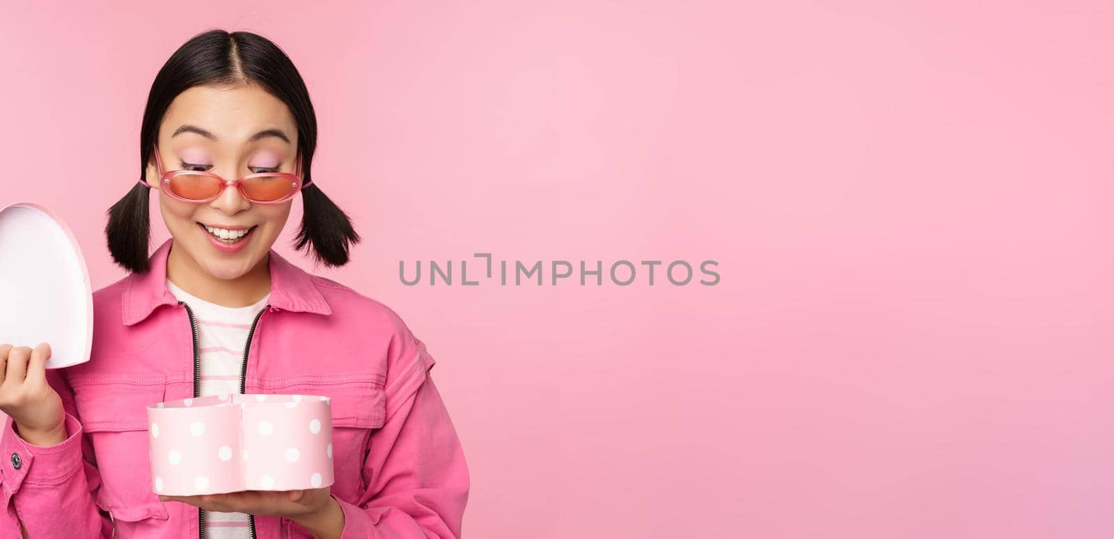Beautiful asian girl opens up heart shaped gift box, looking happy, standing in sunglasses, smiling surprised at camera, pink background.