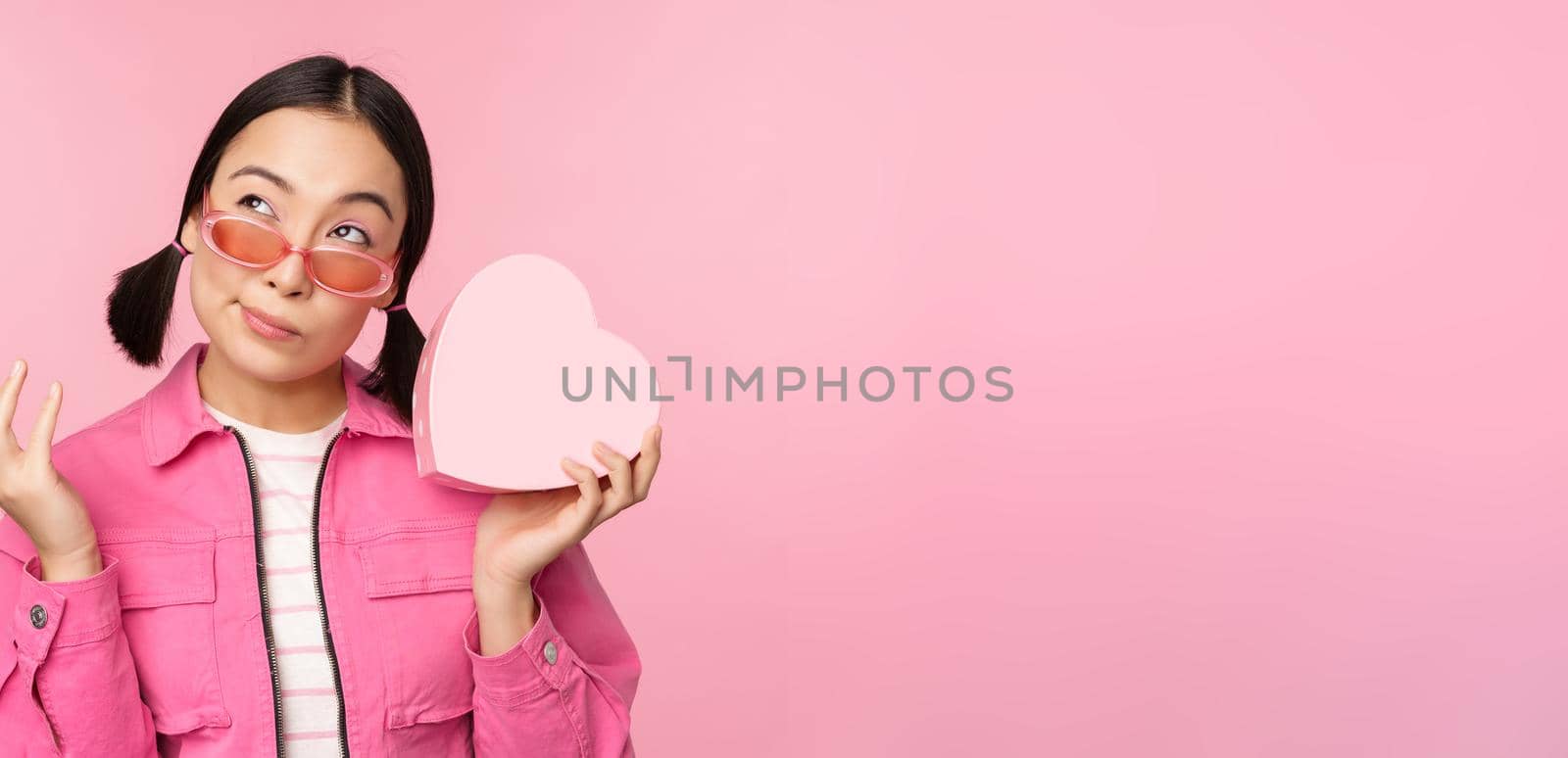 Image of stylish asian girlfriend in sunglasses, guessing whats inside gift box, heart-shaped present, standing over pink background by Benzoix
