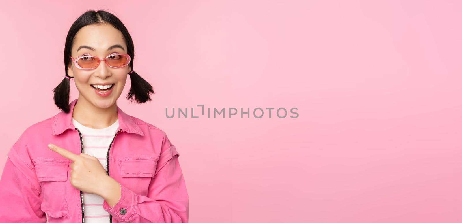 Portrait of smiling asian girl in stylish outfit, sunglasses, pointing finger left, showing advertisement, banner, standing over pink background. Copy space