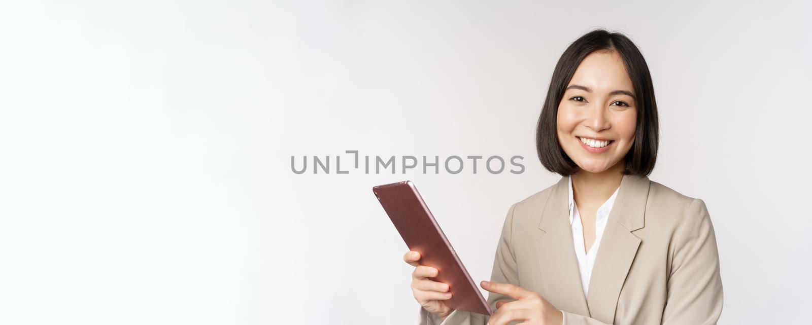Image of asian businesswoman, saleswoman holding digital tablet and smiling, working with gadget, standing in suit over white background.