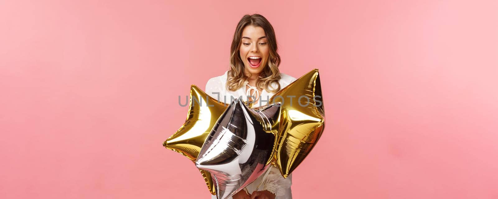 Holidays, celebration and women concept. Portrait of joyful beautiful blond girl in white dress being excited and impressed with cool start-shaped balloons, partying over pink background.