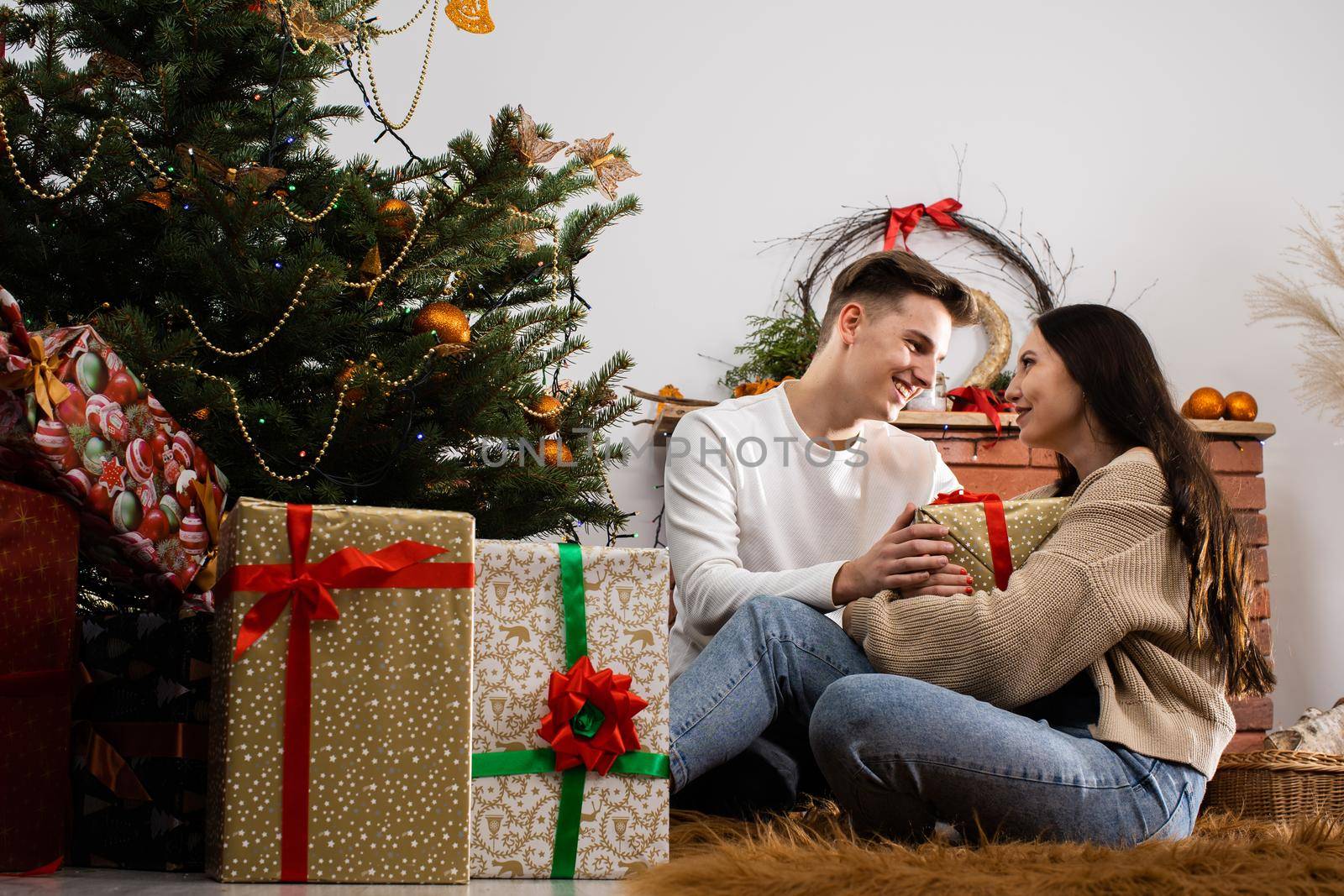 Couple in love sitting by the Christmas tree on the carpet in Christmas. Decorated Christmas tree and fireplace in the guest lounge at home.