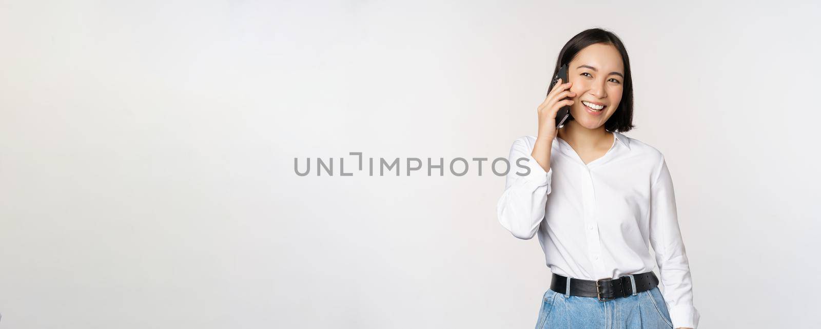 Friendly smiling asian woman talking on phone, girl on call, holding smartphone and laughing, speaking, standing over white background.