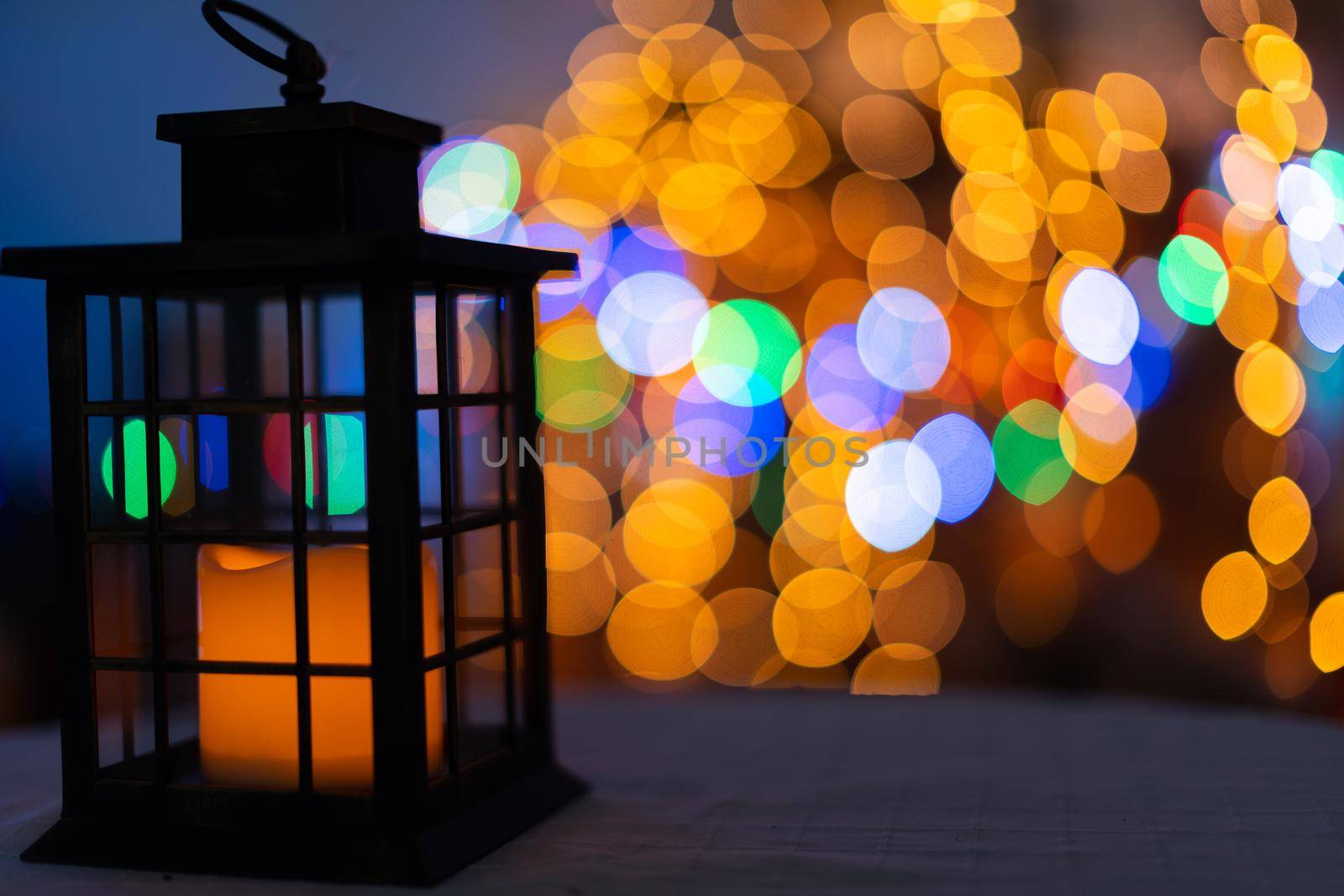 In the night frame a rectangular old hand lantern with a glowing candle in the center. In the distance a blurred background created from the bokeh of glowing Christmas tree lights