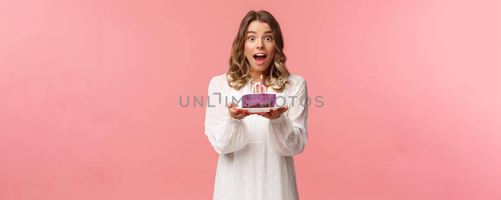 Holidays, spring and party concept. Happy cheerful good-looking blond woman celebrating birthday, holding piece cake with lit candle, making wish, look amused standing pink background.