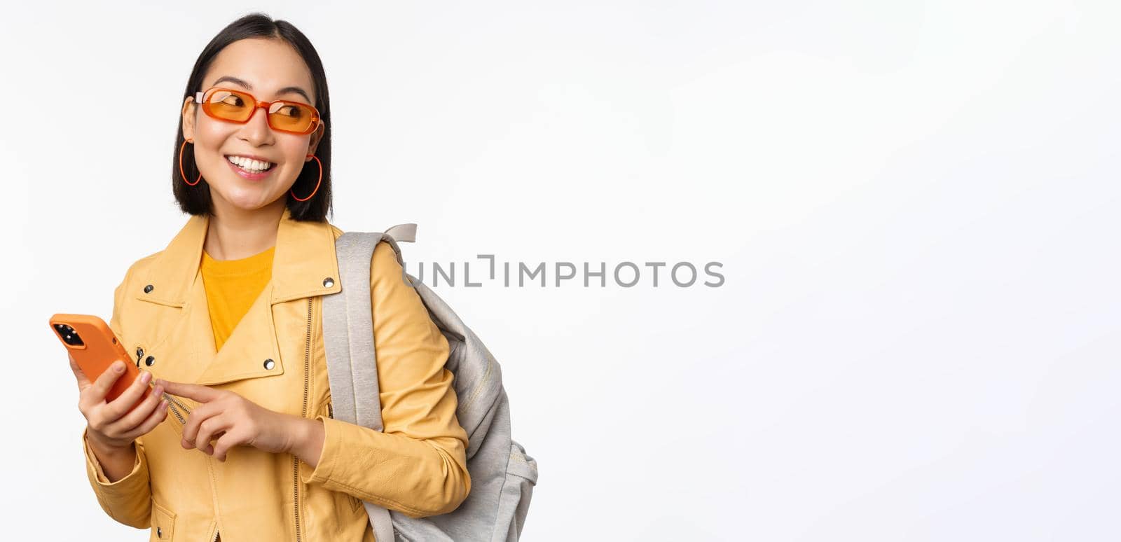 Stylish young asian woman tourist, traveller with backpack and smartphone smiling at camera, posing against white background. Copy space