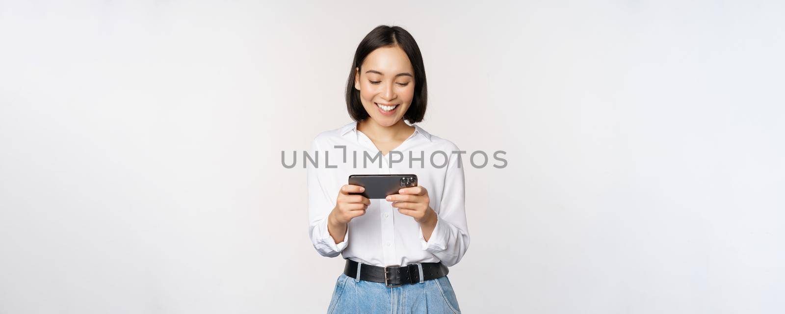 Young korean woman, asian girl playing mobile video game on smartphone, looking at horizontal phone screen, standing over white background.