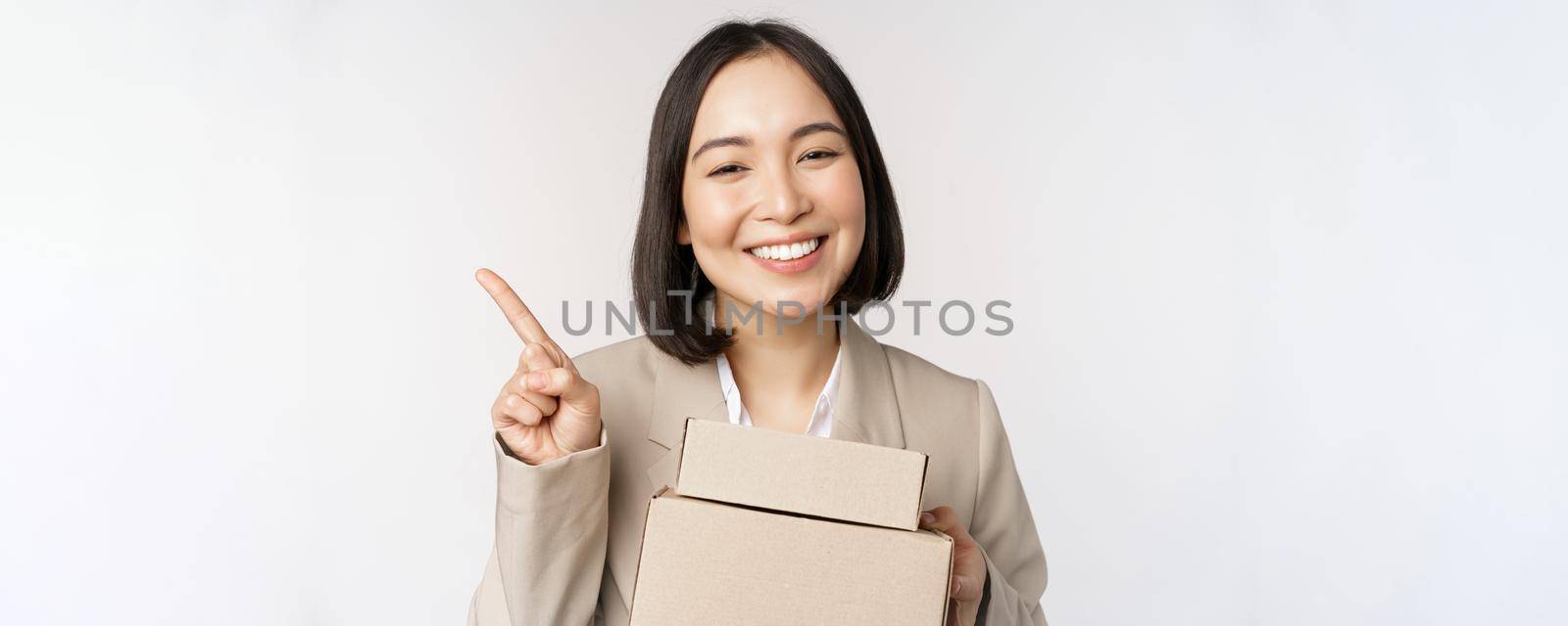Smiling asian saleswoman pointing finger at copy space, holding boxes with delivery order, standing over white background.