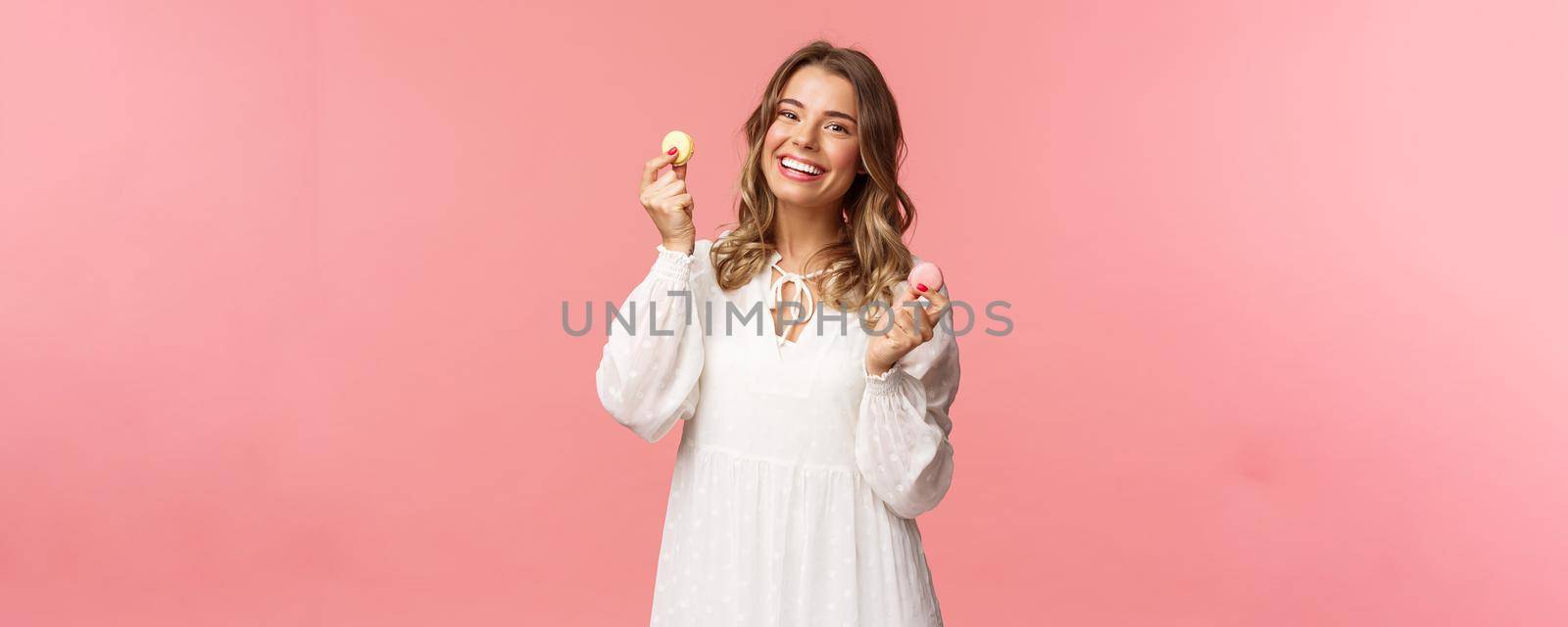 Holidays, spring and party concept. Portrait of cute romantic blond girl in white dress, licking lips as tempting to eat delicious dessert, holding two macarons and look pleased, pink background.
