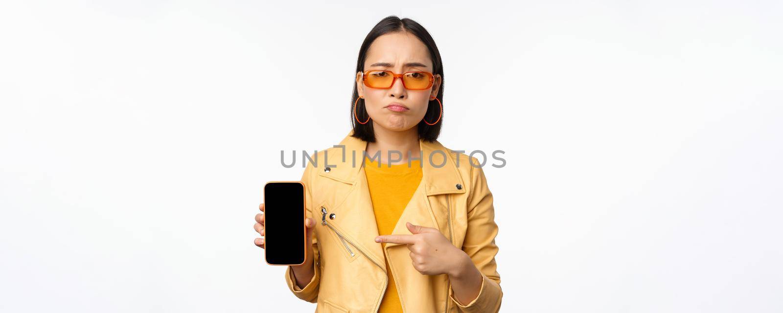 Portrait of sad asian woman in sunglasses, pointing finger at mobile phone app interface, showing smartphone application, standing over white background.
