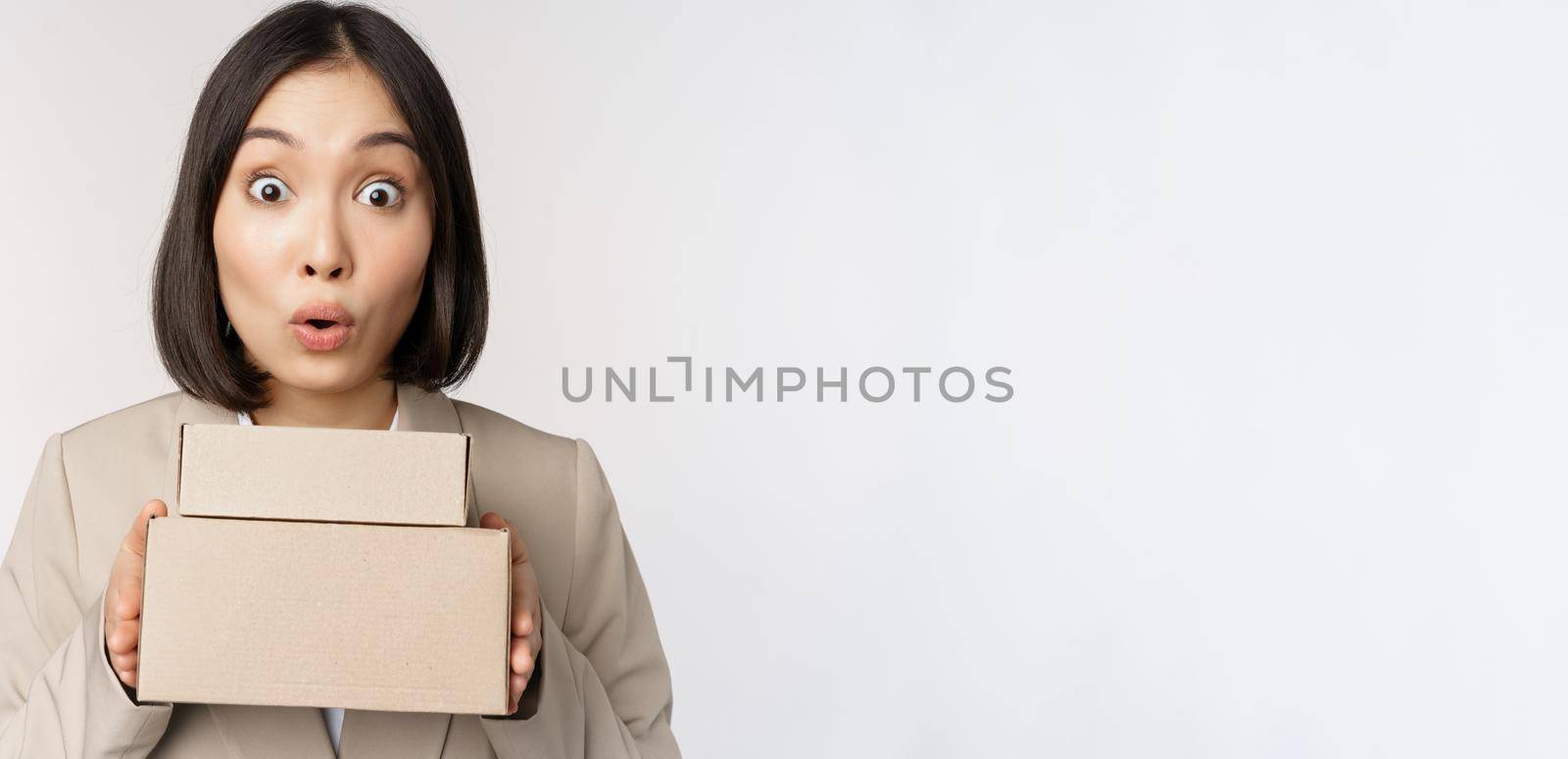 Portrait of asian saleswoman looking surprised, holding boxes, delivery goods, standing amazed in suit against white background.