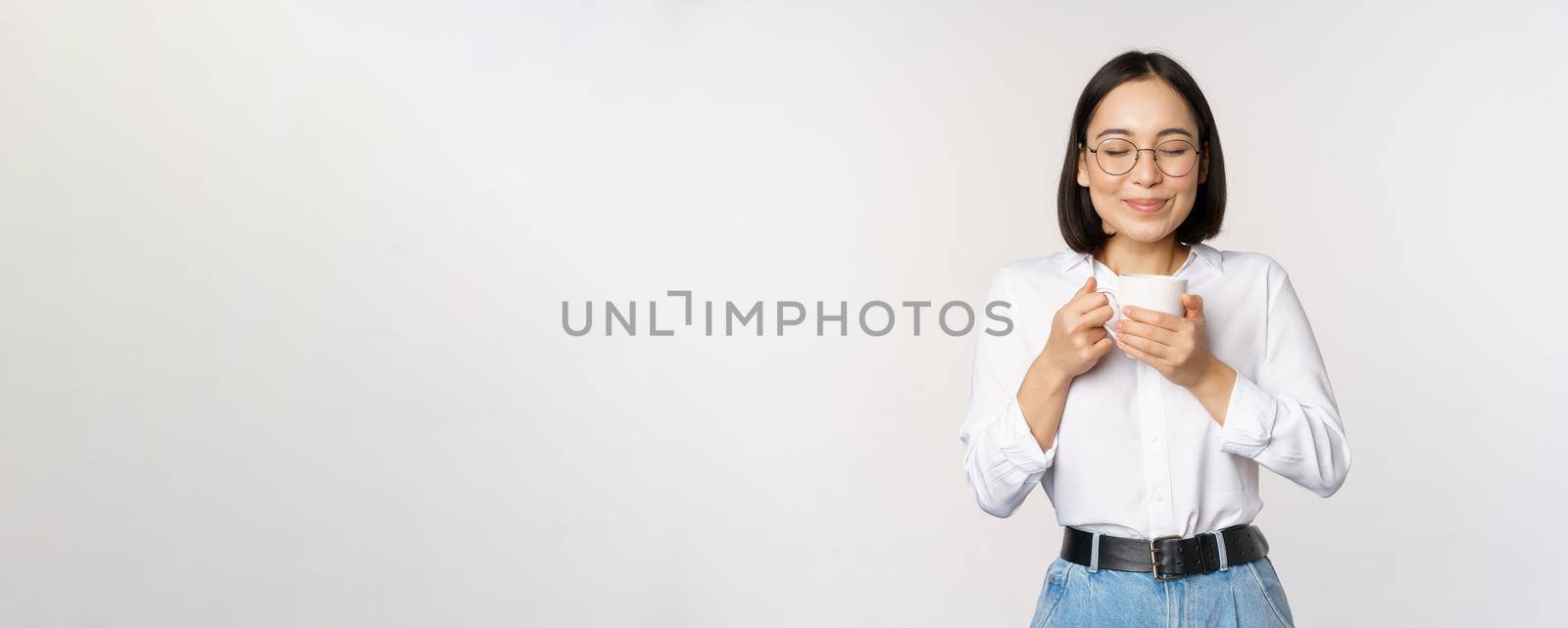 Image of smiling korean woman sniffing, smell coffee in mug, enjoying delicious drink, standing over white background.