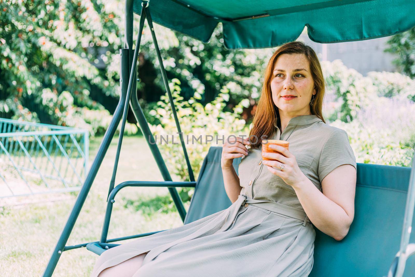 a young girl in the summer drinks hot tea, sitting on a swing, in her yard, tea helps in the heat best of all, by mosfet_ua