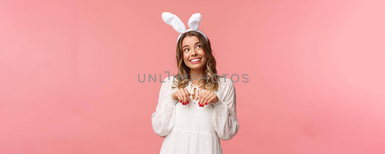 Holidays, spring and party concept. Cute romantic young blond girl imitating bunny, wear white dress lovely rabbit ears, make hand-paws near chest and look dreamy up with daydreaming smile.