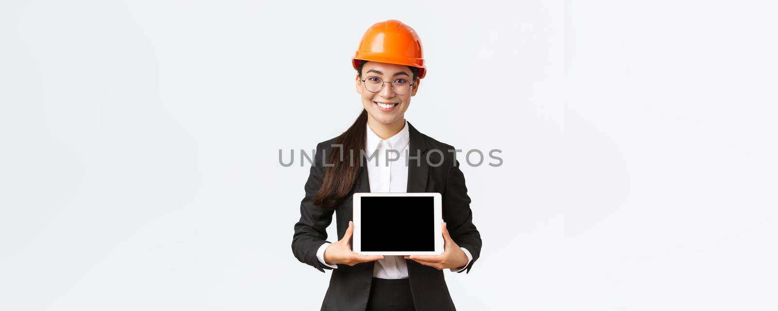 Smiling professional asian female architect introduce her project, engineer showing diagram at digital tablet display, making presentation at enterprise or factory, standing white background.