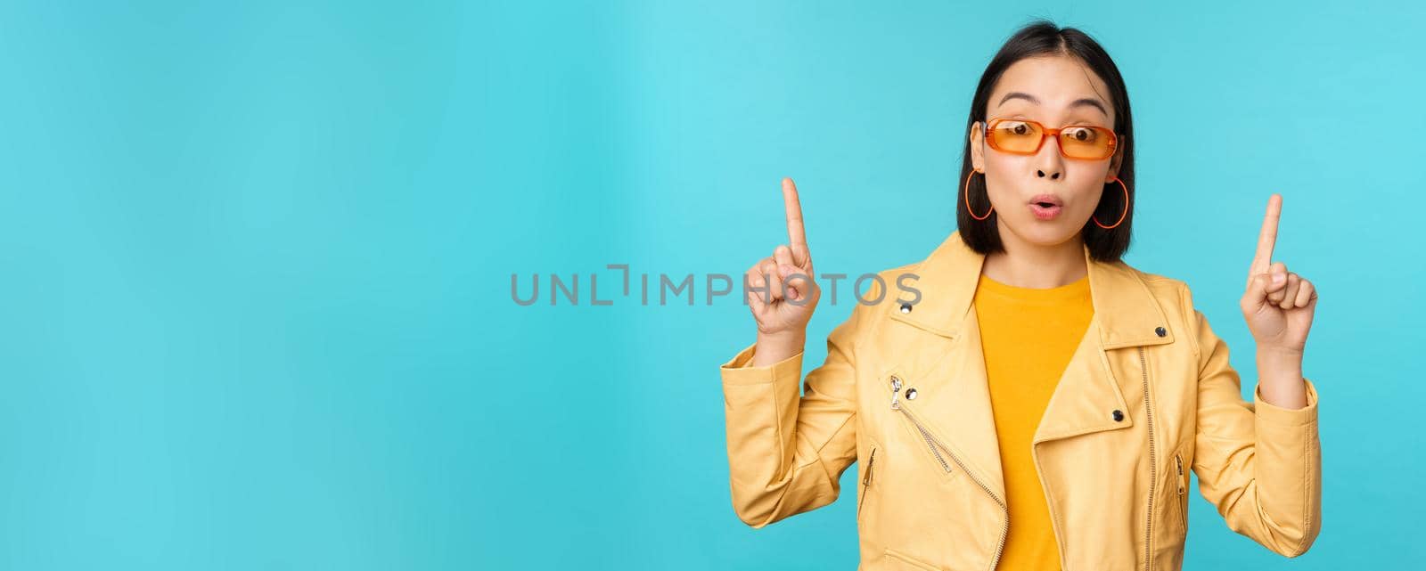 Enthusiastic asian girl in sunglasses, points fingers up, shows banner or logo on top, stands over blue background. Copy space