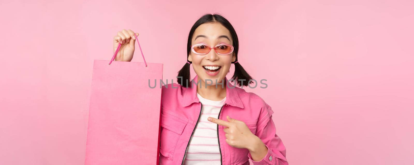 Shopping. Stylish asian girl in sunglasses, showing bag from shop and smiling, recommending sale promo in store, standing over pink background.