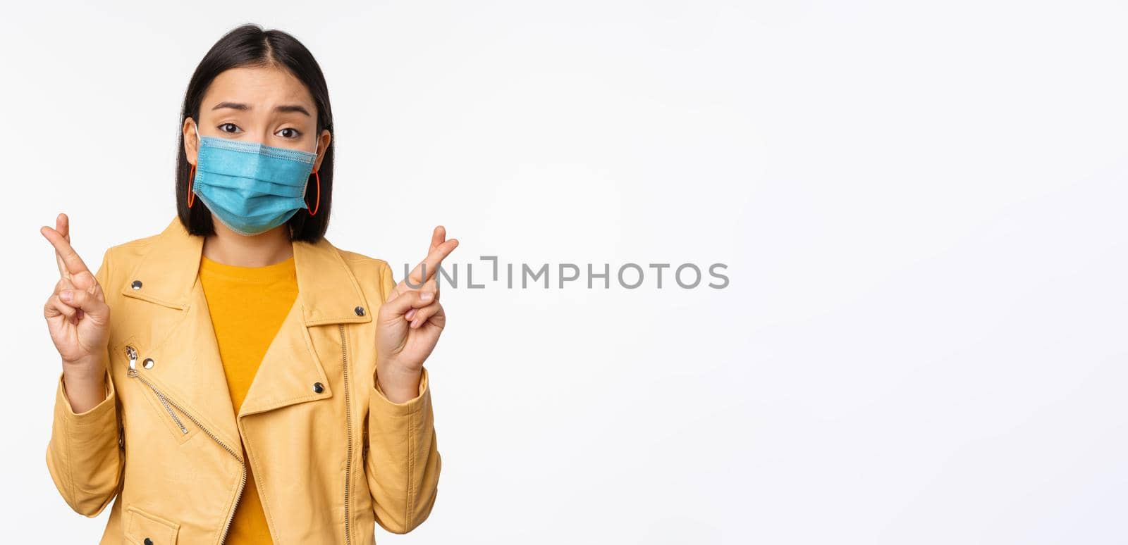 Hopeful asian girl in medical face mask, cross fingers, making wish, hoping, praying for smth, standing with anticipation over white studio background.