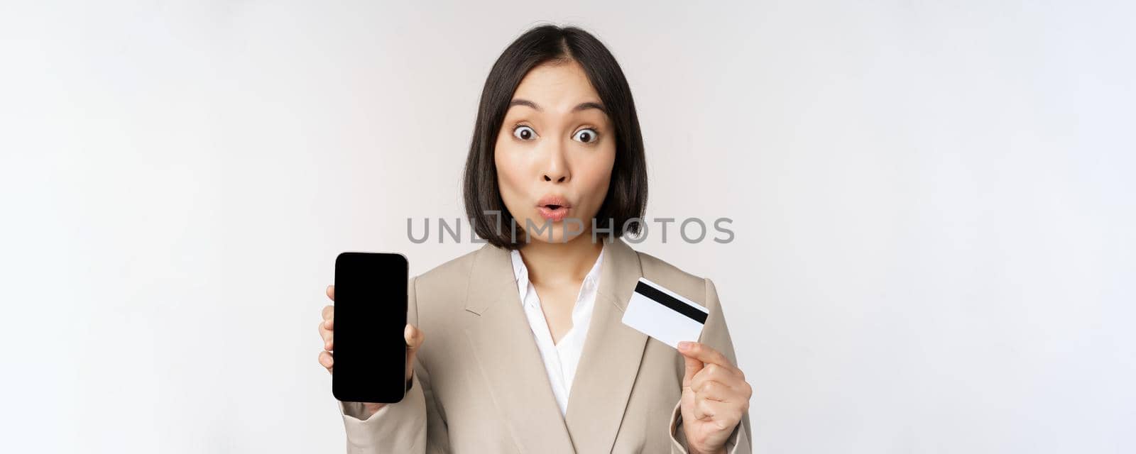 Corporate woman with happy, enthusiastic face, showing credit card and smartphone app screen, standing in suit over white background.