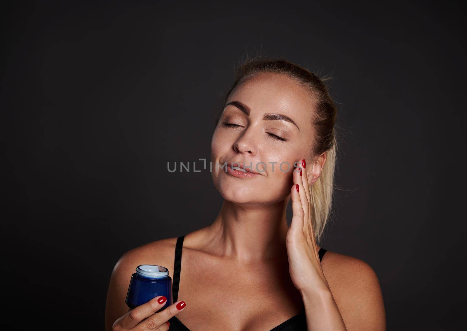 Close-up beauty portrait beautiful Caucasian woman applying moisturizer on face, holding jar of daily smoothing cream or beauty serum, isolated on black background. Skin care, cosmetology, spa concept