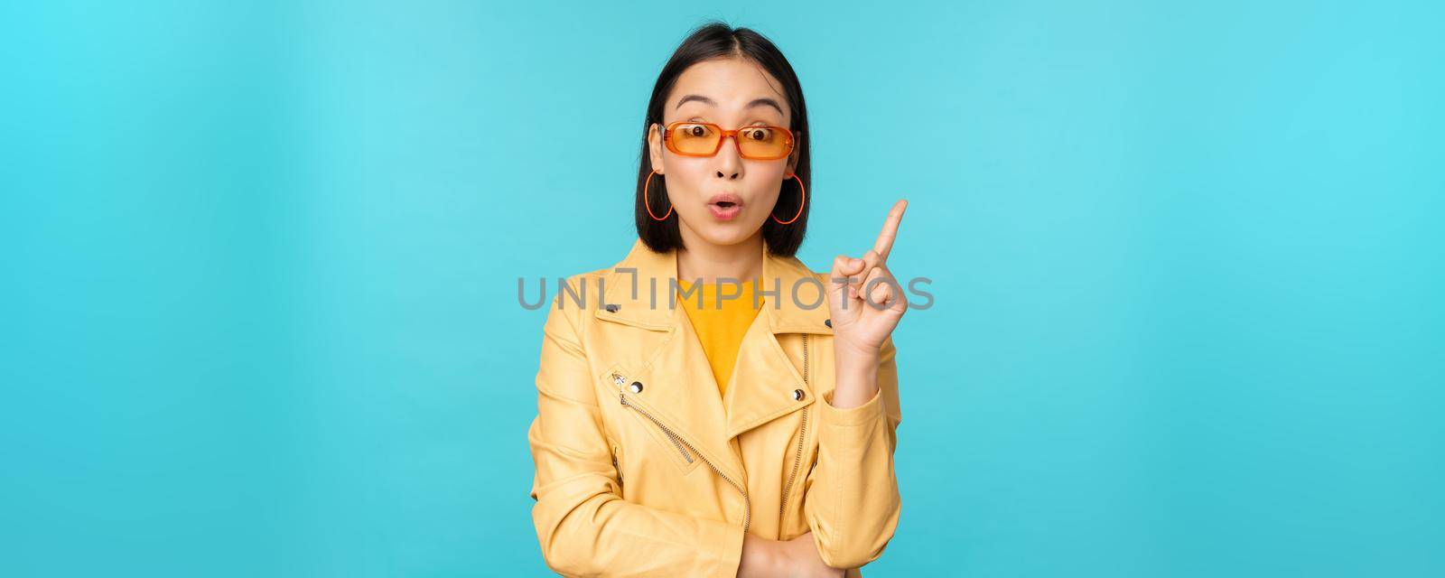 Excited asian girl raising finger, suggesting smth, eureka sign, has plan, standing in sunglasses over blue background.