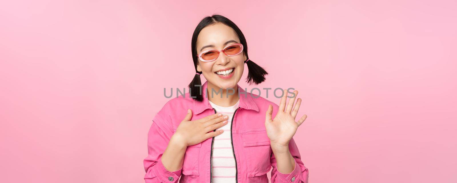 Portrait of friendly asian teen girl in sunglasses saying hello, waves her hand and smiles, greets you, hi gesture, stands over pink background.