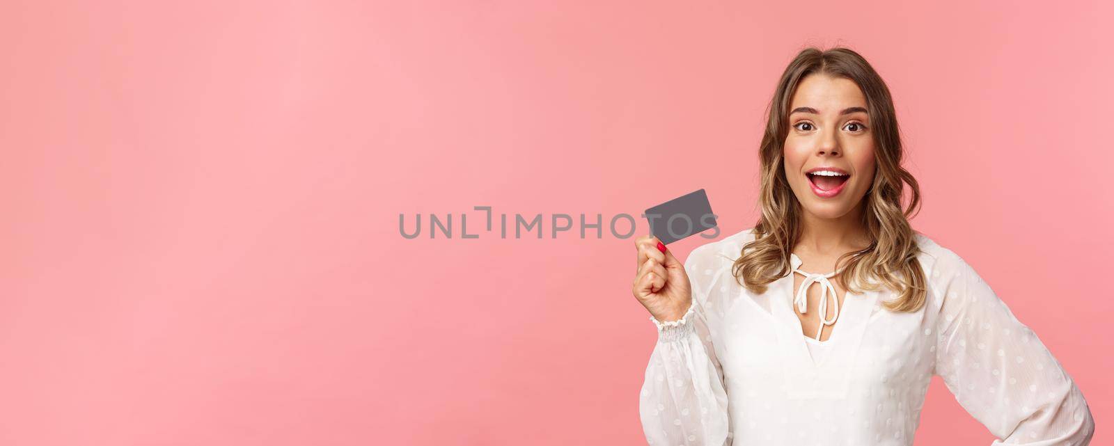 Close-up portrait of excited pretty young blond woman in white dress, holding credit card, showing people how she invests, look camera amused, smiling recommend shopping app, pink background.