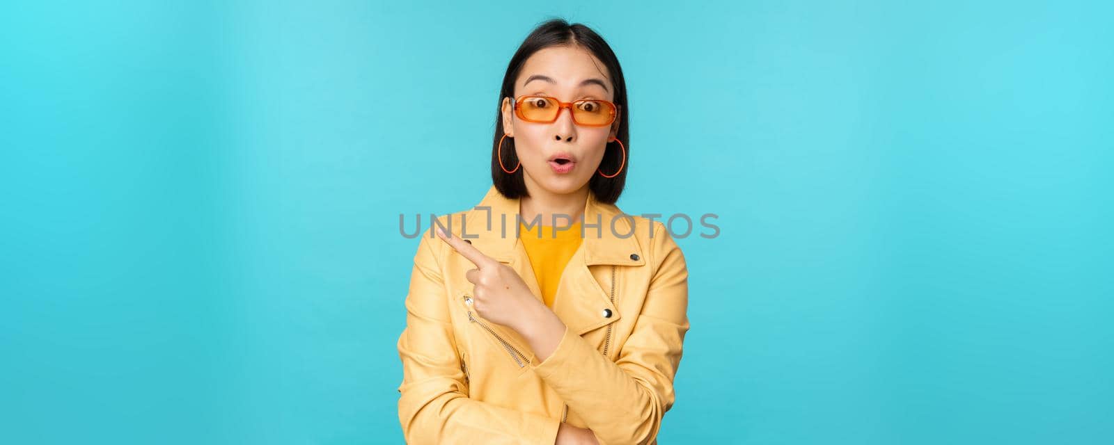 Image of asian woman looks intrigued, asks question about item or store, points finger left with surprised face expression, stands over blue background.
