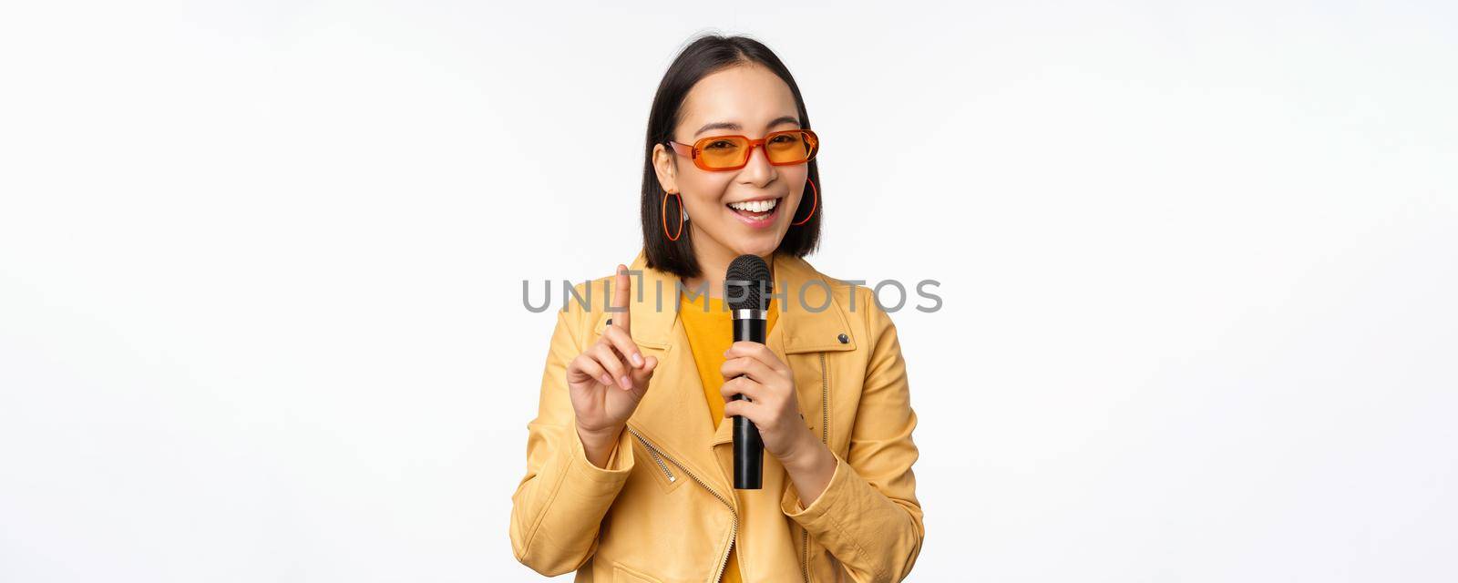 Portrait of beautiful asian woman in sunglasses, stylish girl singing, giving speech with microphone, holding mic and smiling, standing over white background. Copy space