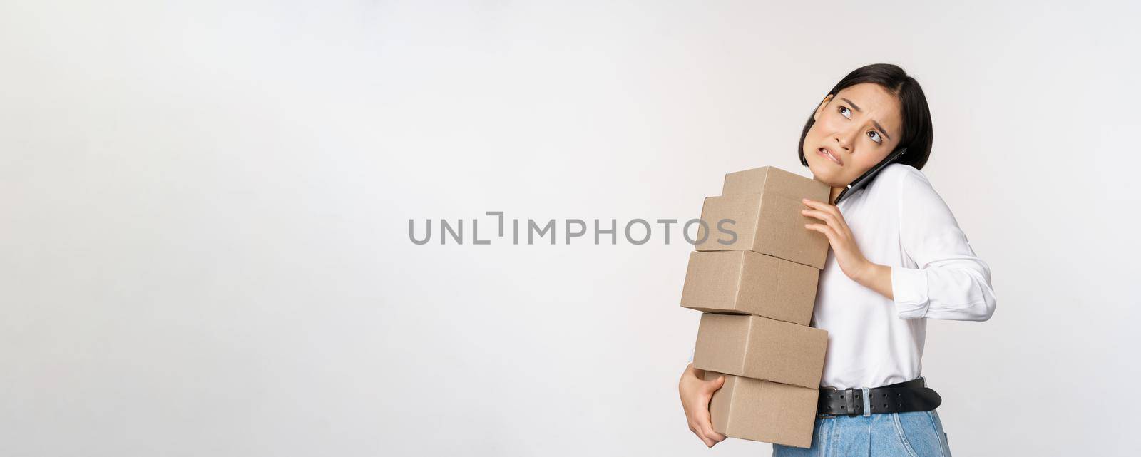 Young asian businesswoman answer phone call, talking on mobile while carrying pile of boxes with orders, standing over white background.