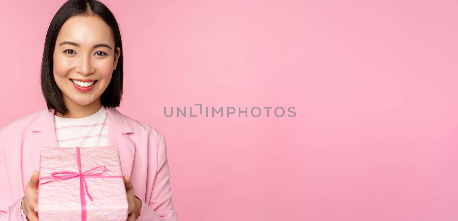 Smiling asian businesswoman in suit, giving you gift in wrapped box, standing over pink background. Copy space