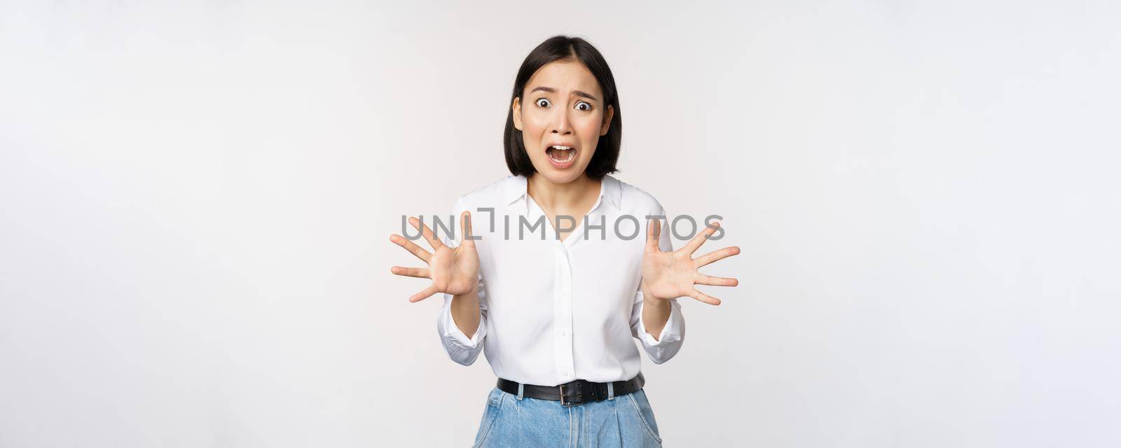 Asian woman looks at camera and screams in panic. Young korean girl looking anxious, panicking, shaking hands and shouting, standing over white background.