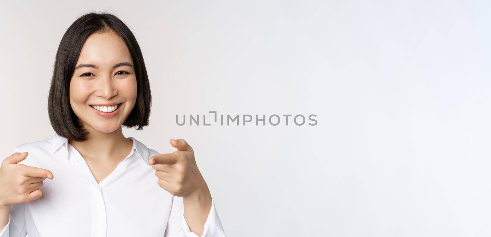 Close up of enthusiastic young woman smiling, pointing fingers at camera, choosing you, inviting and recruiting people, standing over white background.