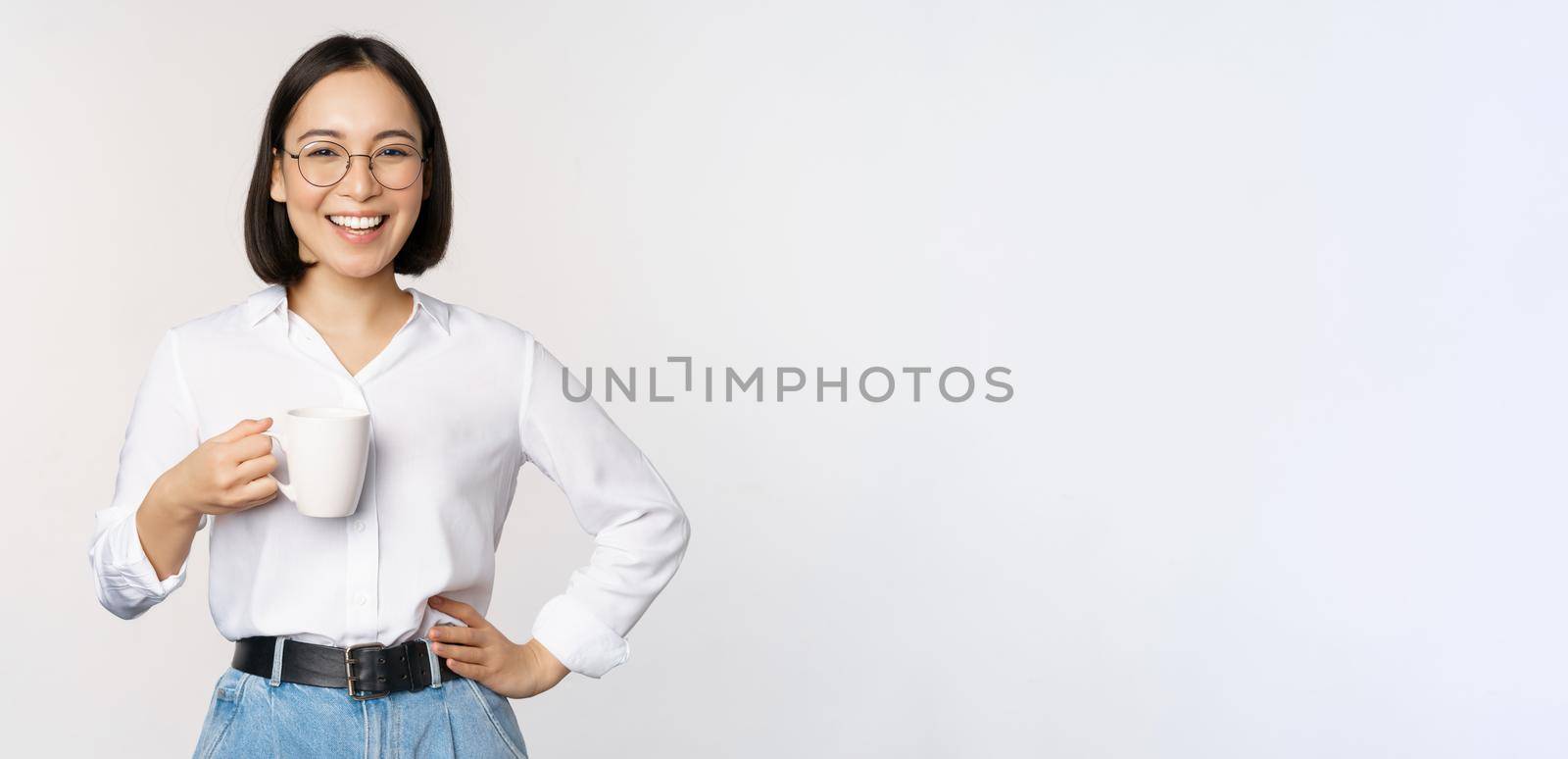 Happy young energetic asian woman smiling, drinking, holding cup mug of coffee, standing confident against white background.