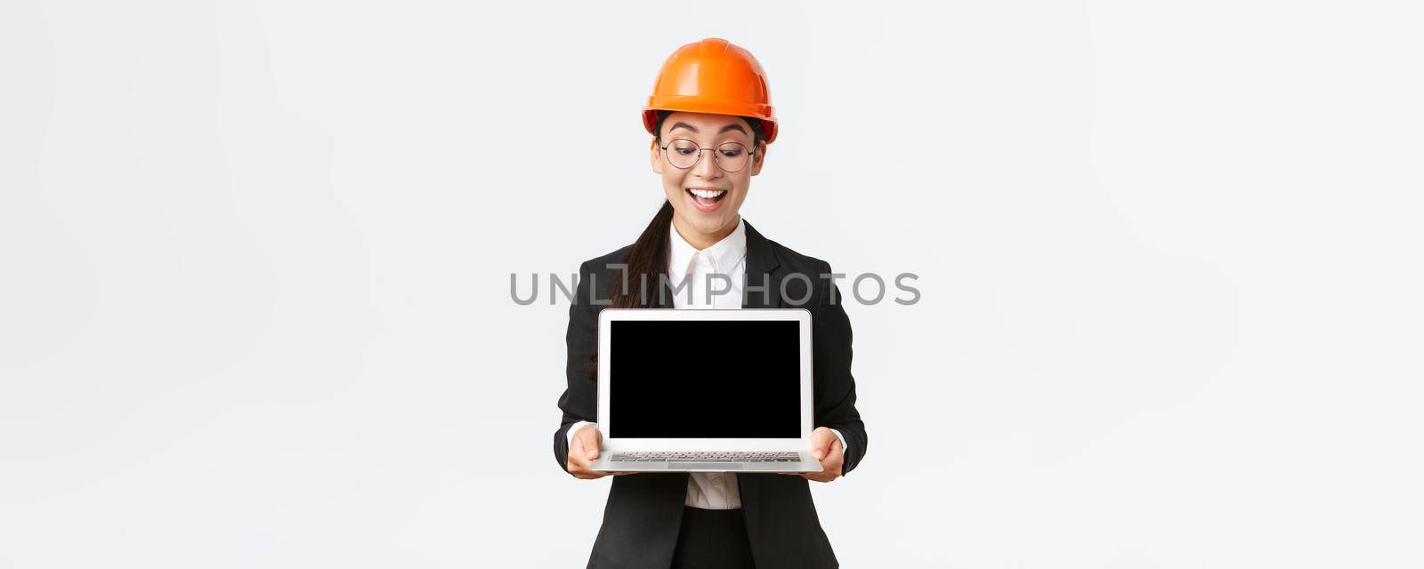 Impressed happy asian female chief engineer showing great results of enterprise to investors, standing in safety helmet and business suit, holding laptop and look pleased at computer screen.