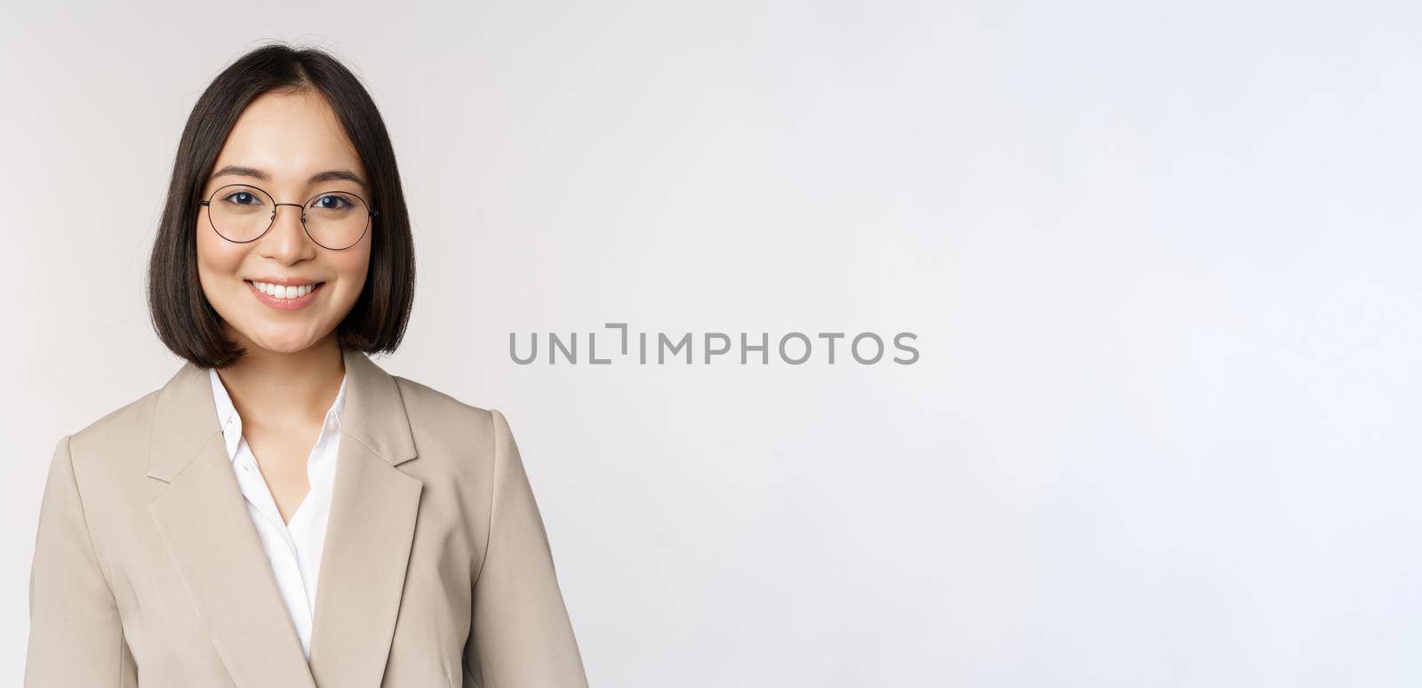 Portrait of young asian saleswoman in glasses, wearing beige suit, smiling and looking confident at camera, white background.