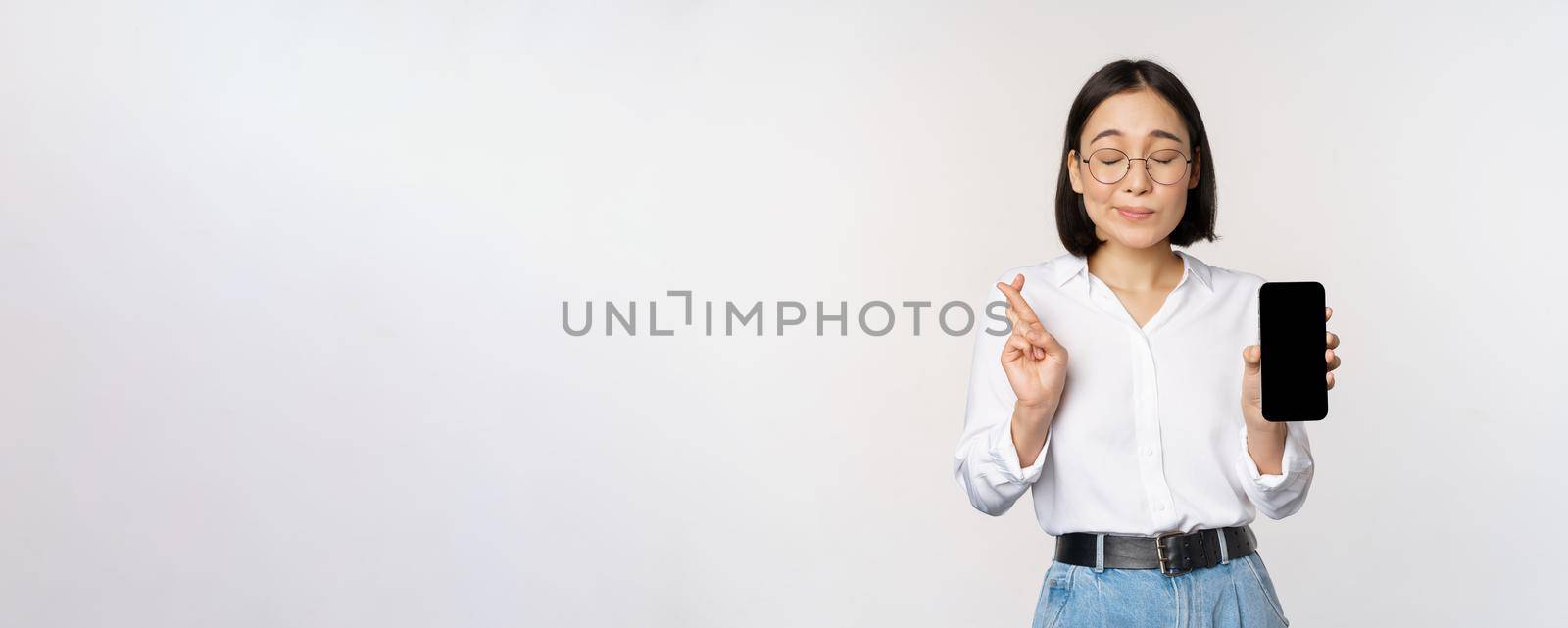 Hopeful young asian woman, showing mobile phone screen, app interface and fingers crossed, hoping for smth, making wish or paying, standing over white background.