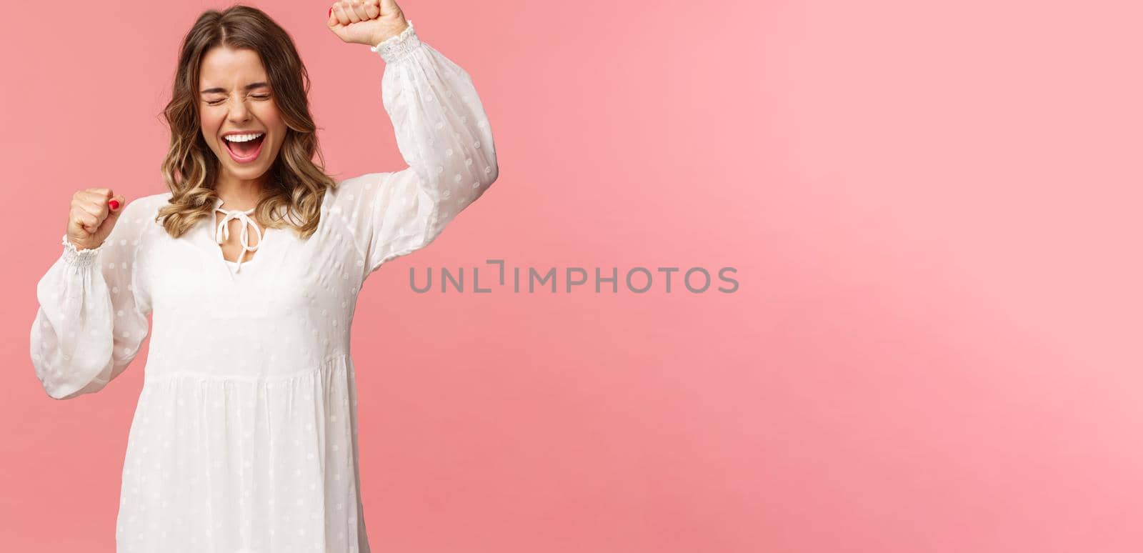 Portrait of beautiful tender young blond girl in white dress, raising hands up in hooray, yes or victory, shouting relieved, scream from happiness, triumphing, winning prize, become champion.